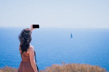 A young girl in a blue red dress takes pictures on her phone of the sea and a small sailboat