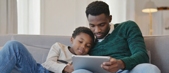 Father-Son Bonding: A tender moment unfolds as a father and son share a tablet on a comfortable sofa, the soft lighting and intimate atmosphere emphasizing their close relationship.