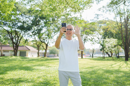 Happy senior asian man waving while taking selfie with smartphone outdoor in green park. He enjoys modern technology on sunny day