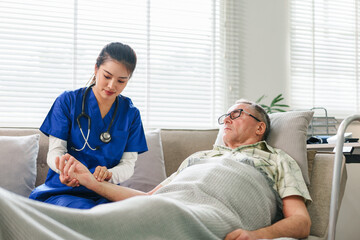 Obraz premium Female nurse checking pulse of senior male patient lying on sofa during home care visit, caregiver monitoring elderly man's health