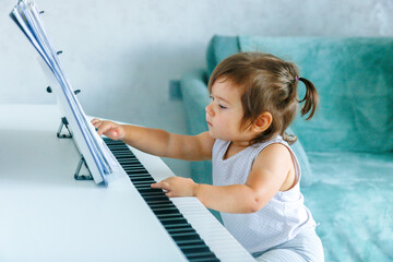 A one-year-old girl at the piano looks with interest at the sheet music on the music stand