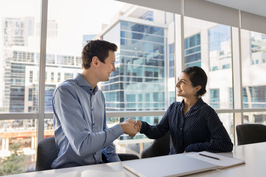 Diverse smiling millennial coworkers handshake sitting at office work desk