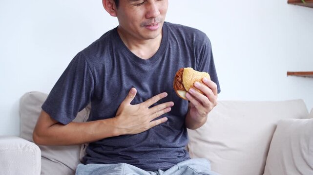 Asian man eating a burger while experiencing digestive system problems such as acid reflux and GERD. He suffering from discomfort, stomachache, and heartburn after eating unhealthy junk food. 