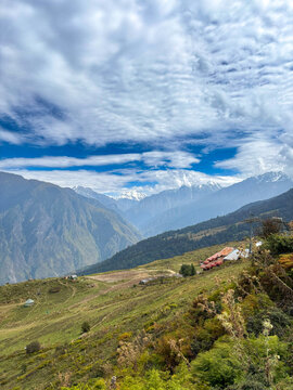 Auli, Uttarakhand, India - Panoramic View of Snow-Capped Nanda Devi Peak and Himalayan Mountain Range Under Clear Blue Skies with Lush Green Meadows and Traditional Hill Village Structures