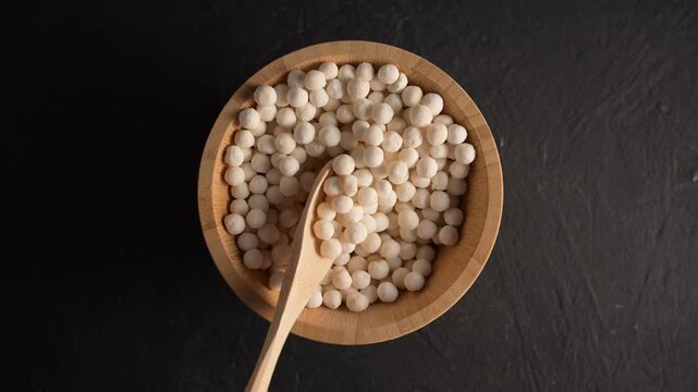 Organic tapioca pearls in wooden bamboo bowl on black table top view. Spoon scooping white dry tapioca pearls, cooking ingredient, gluten-free food, healthy eating.