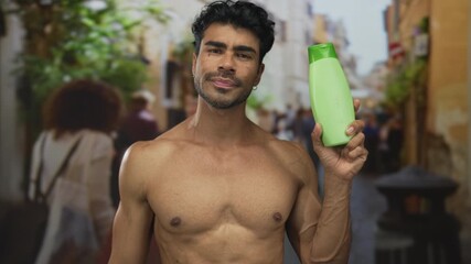 Shirtless hispanic man holds green bottle near his bare chest on a bustling city street filled with blurred pedestrians; confidence vitality wellness energy.
