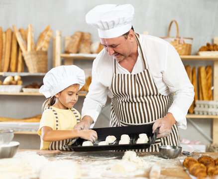 Little girl in striped apron and white chefs hat near kitchen table puts croissants on baking sheet