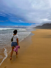 Young Woman Walking on Cofete Beach, Fuerteventura, with Backpack on a Sunny Cloudy Day