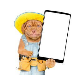 Smiling Mastiff puppy wearing summer hat and denim overalls with tool belt showing big smartphone with white blank screen in it paw. Isolated on white background