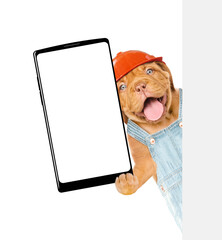 Happy Mastiff puppy wearing hard hat and denim overalls holding big smartphone with blank screen in it paw behind blank wihte banner. Empty space for text. Isolated on white background