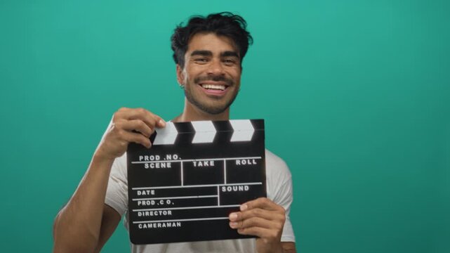 Hispanic man in white t shirt holding film clapboard against vibrant green studio wall with confident posture; creativity imagination inspiration innovation.