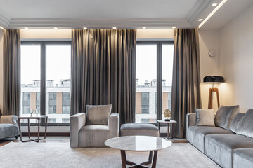 luxurious interior of large apartment with large windows, gray armchairs and sofas. light carpet on  floor and a coffee table. A lamp is shining in the corner. the atmosphere of coziness and comfort