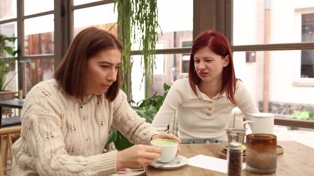 Two young women sit in a cafe one tastes matcha tea and makes a disgusted face while the other reacts with surprise and discomfort funny moment of trying a new drink with unpleasant flavor reaction