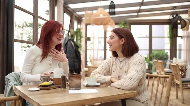 Two friends sit in a cozy cafe laughing and chatting warmly one tells a funny story with open gestures while the other listens with a bright smile warm daylight relaxed friendly atmosphere
