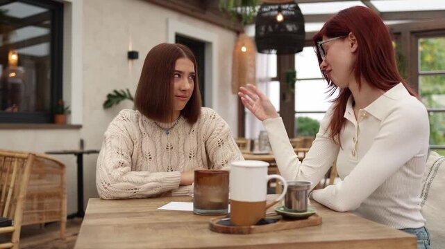 Two students sit in a cozy cafe one teasing the other about her old sweater while the other reacts with quiet discomfort warm daylight relaxed setting but subtle tension between them