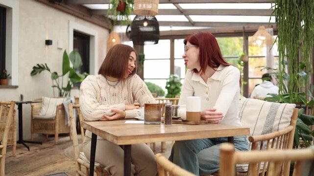 Two sisters sit in a cozy cafe playfully sticking their tongues out at each other sharing a lighthearted teasing moment warm daylight relaxed friendly atmosphere casual connection
