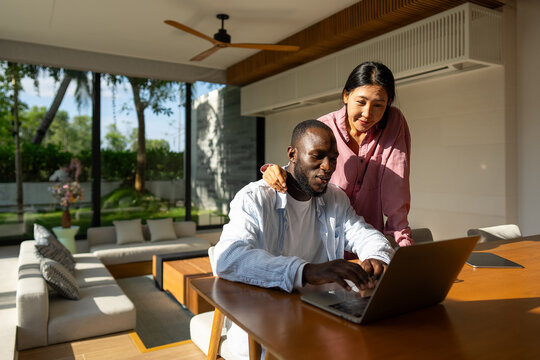 Multinational couple enjoying their morning routine, smiling and using laptop and tablet at home
