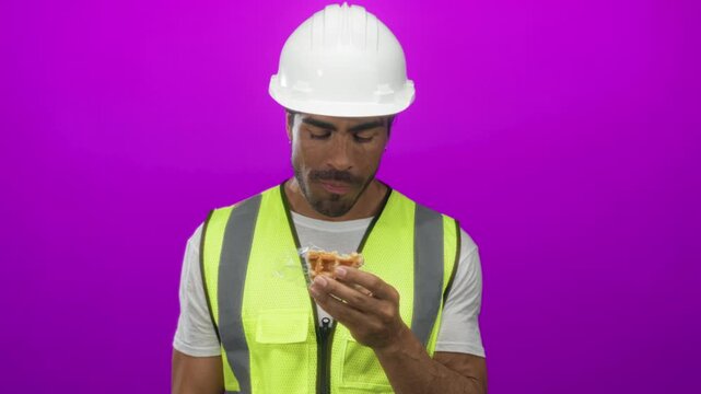 Hispanic man bites waffle while holding wrapper in purple studio; hunger break nourishment satisfaction.