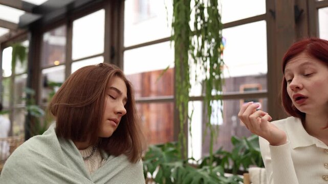 One student exhausted after classes sits wrapped in a blanket and has fallen asleep mid conversation while her friend continues talking unaware of her tired state calm everyday moment