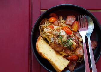 Top view food of spaghetti bacon with fried garlic and tomato serve on black dish with toast on red wooden table.