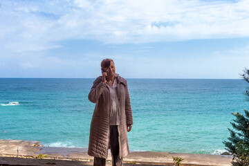 Fototapeta premium Woman talking on smartphone by sea. Communication technology concept with mature female. Travel destination for vacation.