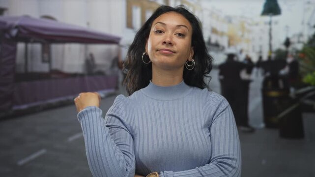Woman with palm down dismissive gesture and crossed arms wearing a light blue ribbed sweater on street market backdrop; confidence.