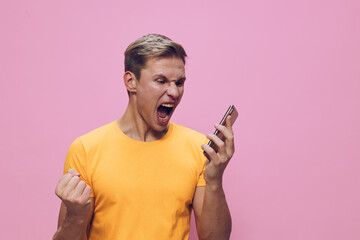 Smiling young man in yellow t-shirt expressing excitement and joy while looking at mobile phone isolated on pink background. People lifestyle concept, success, celebration, happiness, positive emotion