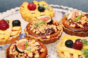 Fruit tartlets with crumble topping, figs, raspberries, and blueberries on silver tray. Homemade mini desserts with sweet filling and mint decoration. Delicious bakery treats. Glass tray presentation.