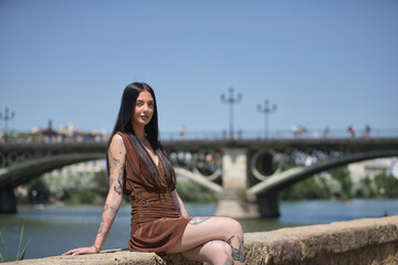 Young woman, brunette, tall, thin and very tattooed, with brown dress, posing looking at the camera sitting by the river with the Triana bridge in the background. Concept of youth, beauty.
