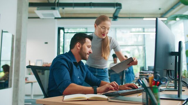 Smiling woman standing beside seated male coworker holding clipboard with data. Man typing on keyboard checking information online. Colleagues verifying report together in bright office space.