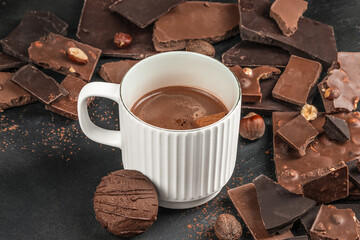 Close-up of white ceramic mug with hot chocolate and cookie surrounded by dark and milk chocolate pieces with hazelnuts on black textured background, cozy winter drink concept, top view copy space