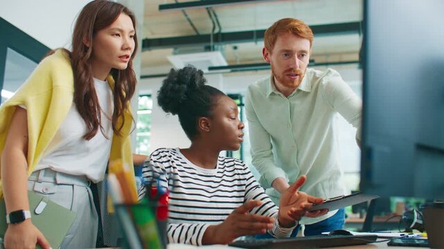 Team of diverse coworkers gathered around desk discussing error on computer screen. Red-haired man pointing while women listening attentively. Group solving work issue in collaborative office.