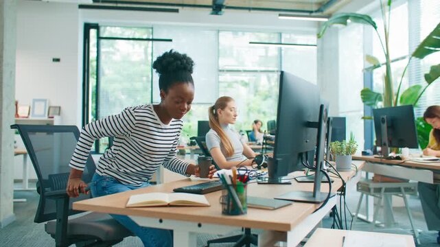 Pretty African American girl in striped shirt arriving at workplace holding takeaway coffee. Female employee sitting down at desk preparing to start workday in bright shared office.