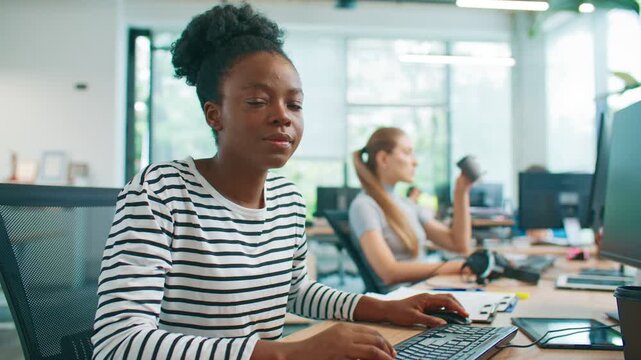Beautiful African American woman in striped shirt smiling and looking directly at camera while sitting at desk. Female employee distracted from tasks on table in bright shared office workspace.