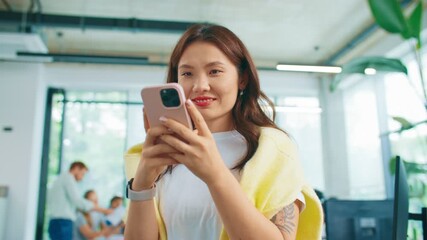 Smiling woman standing in bright office holding smartphone and typing message. Engaging in online chat or social media conversation. Relaxed atmosphere with colleagues working in background.