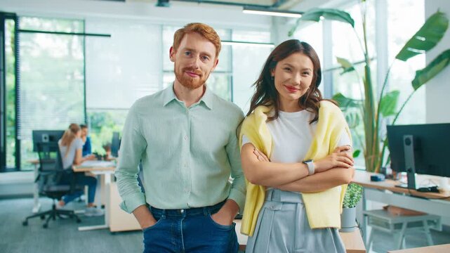 Two colleagues standing side by side in bright workspace while smiling confidently. Creative team working in background. Office environment filled with light and modern equipment. Posing together.