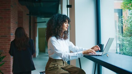 Curly-haired Caucasian woman sitting near window working on laptop focused on screen. Wearing white shirt and brown trousers. Modern corridor with brick walls and plants. Blurred colleague passing by. - Powered by Adobe