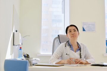 Obraz premium Young female Asian physician with stethoscope sitting at working table in consulting room and looking into the distance
