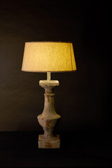 A rustic table lamp with a glowing lampshade stands against a dark background. Its vintage-style base shows distressed textures, creating a warm, cozy, and old-world ambiance.