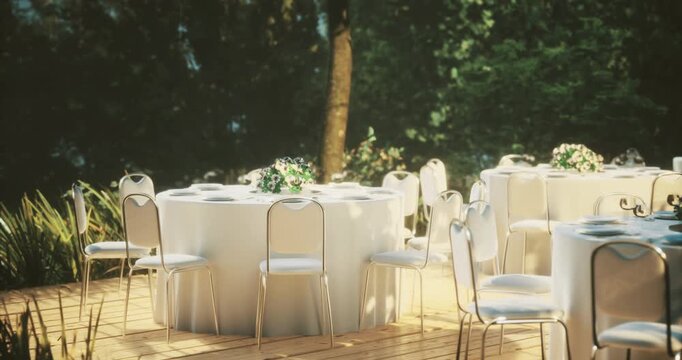 An outdoor venue is prepared for a wedding celebration, showcasing round white tables adorned with small floral centerpieces. Natural greenery enhances the romantic atmosphere.
