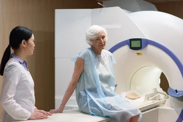 Fotobehang Beren Senior Caucasian woman sitting on bed of MRI machine after being scanned, young Asian female nurse assisting her  © pressmaster