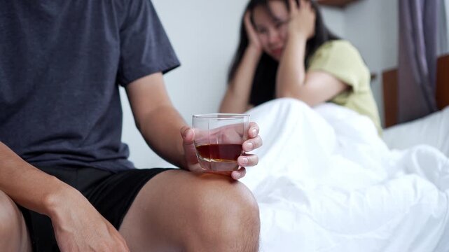 Asian man drinking alcohol in bed while his bored and frustrated wife sits beside him feeling ignored. Scene shows alcohol addiction, relationship problems, family conflict, stress in marriage.
