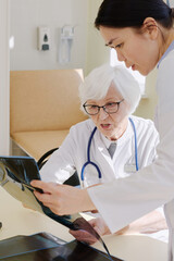 Fototapeta premium Mature female physician analyzing conditions of patient by looking at his X-ray after MRI scan and consulting with younger assistant