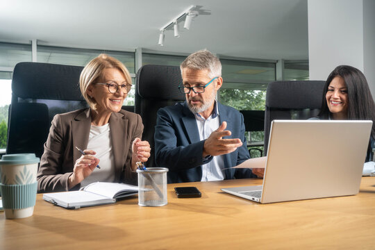 Three busy happy middle aged professional business man and two women executive leaders team using laptop working on computer at work desk having conversation on financial project at meeting in office.