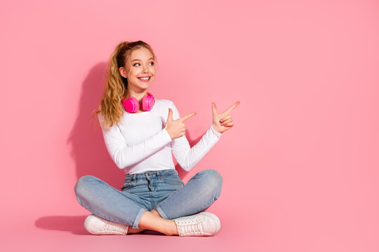 Cheerful young woman with headphones sitting cross legged on pink background pointing in excitement with a joyful gesture
