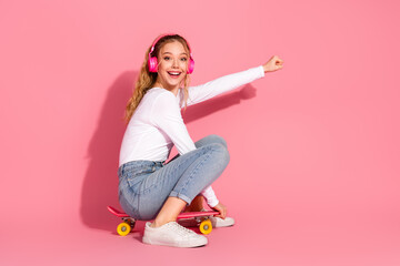 Young girl sitting on a pink skateboard wearing headphones, smiling brightly against a vibrant pink...