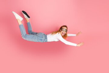 Young girl flying playfully in mid-air while smiling over a pink background wearing casual fashion...