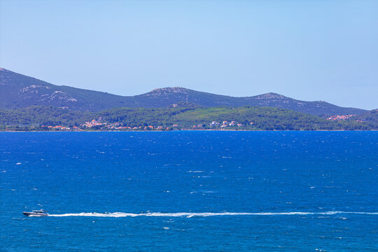 Motorboat speeds across the blue Adriatic Sea near Zadar, Croatia, leaving a foamy trail behind. Ugljan Island appears in the distance with green hills and coastal villages under a clear summer sky