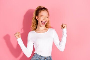 Cheerful young woman celebrating success with raised fists on a pink background, showcasing joy and...