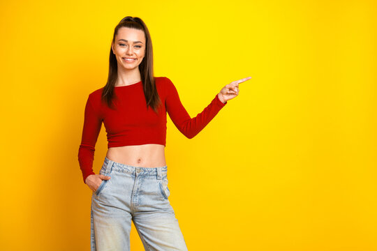 Fototapeta Young brunette woman in a red crop top and denim jeans smiles brightly and points to the right against a bold yellow background for fashion lifestyle advertising